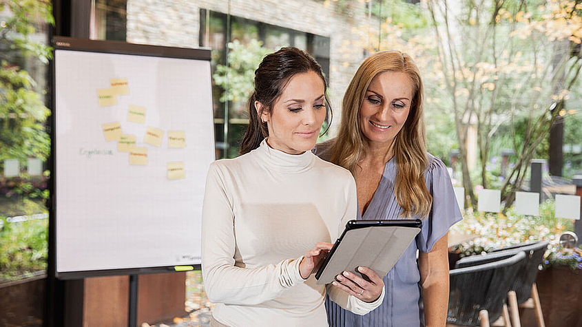 Zwei Mitarbeiterinnen arbeiten an einem Tablet und Whiteboard im Hintergrund